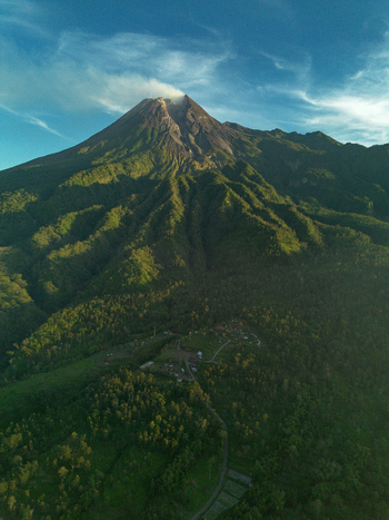 Aerial view of Mount Merapi volcano with smoke rising from the crater surrounded by lush green forested slopes and small village settlements at the base in Indonesia.