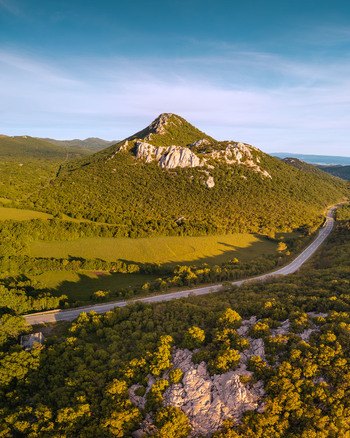 Aerial view of a prominent rocky peak surrounded by lush green forests and a winding road under a clear blue sky in Rijeka Primorje Gorski Kotar Croatia. Aerial view of a prominent rocky peak surrounded by lush green forests and a winding road under a clear blue sky in Rijeka Primorje Gorski Kotar Croatia.