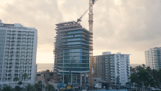 Aerial view of a modern high rise building under construction with a large yellow tower crane surrounded by coastal hotels and palm trees in Miami Beach Florida United States. Aerial view of a modern high rise building under construction with a large yellow tower crane surrounded by coastal hotels and palm trees in Miami Beach Florida United States.