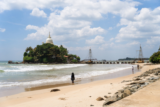 View of Parevi Duwa Temple on a small island connected by a bridge to the sandy beach with people walking along the shore in Matara Southern Province Sri Lanka.