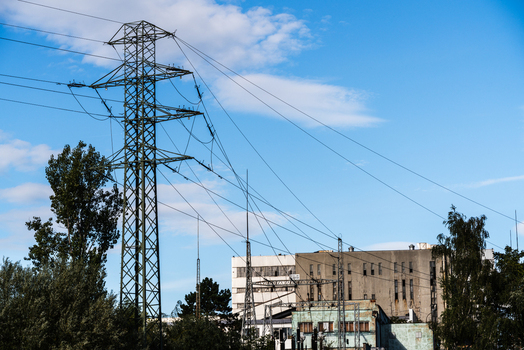 View of a tall metal power transmission tower with electrical lines stretching across a clear blue sky set against an industrial building and surrounding trees in Poland.