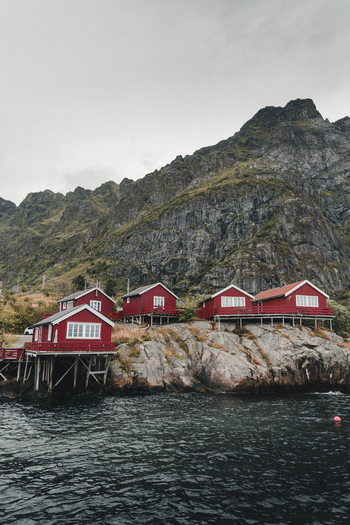 A Nordland Norway 04 September 2025 View of traditional red rorbuer cabins perched on rocky cliffs above the water with a dramatic mountain backdrop in the village of A.