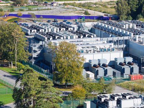 Aerial view of a large data center facility with numerous cooling units on the roof surrounded by trees and parking areas in the United Kingdom.