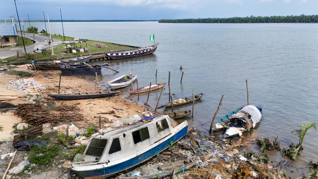 Aerial view of wooden boats and a small motorboat docked along the sandy riverbank with lush greenery and a road nearby in Minima Rivers Nigeria.