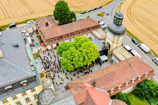 Aerial view of Schloss Jagersburg featuring a courtyard filled with people historic red tiled roofs and a prominent tower surrounded by green trees in Eggolsheim Bayern Germany. Aerial view of Schloss Jagersburg featuring a courtyard filled with people historic red tiled roofs and a prominent tower surrounded by green trees in Eggolsheim Bayern Germany.