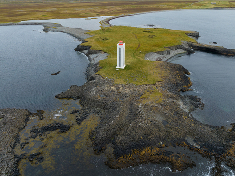 Aerial view of a solitary white lighthouse with a red top standing sentinel on a rocky outcrop amidst the dark swirling waters K&aacute;lfshamarsv&iacute;k Iceland.
