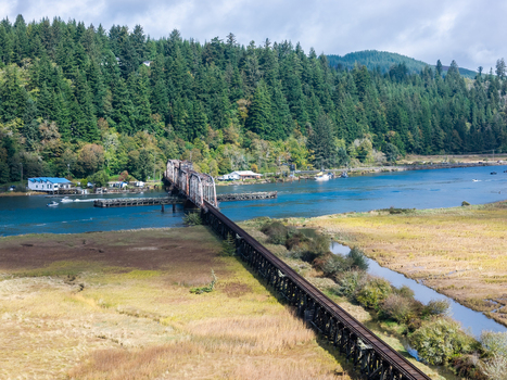 Aerial view of the historic Siuslaw River Train Bridge spanning the river with surrounding lush pine forests and marshy wetlands in Florence Oregon United States.