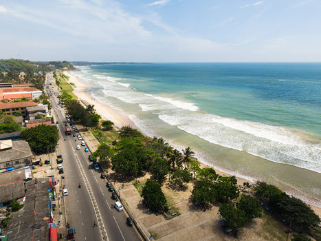 Aerial view of the sandy coastline featuring rolling waves a busy coastal road and lush tropical trees in Matara Southern Province Sri Lanka. Aerial view of the sandy coastline featuring rolling waves a busy coastal road and lush tropical trees in Matara Southern Province Sri Lanka.