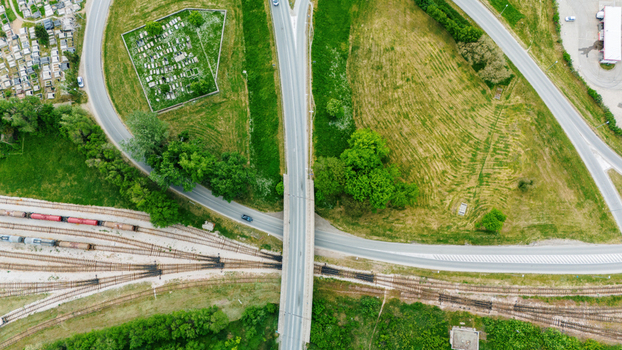 Aerial view of a road bridge crossing over railway tracks with freight trains a cemetery and lush green fields under bright daylight in Sabac Vojvodina Serbia.