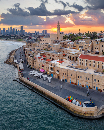 Aerial view of the historic Old port of Jaffa with stone buildings colorful boats at the quay and the modern Tel Aviv skyline under a sunset sky in Tel Aviv Yafo Tel Aviv District Israel.