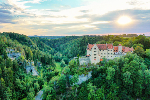 Aerial view of Burg Rabenstein perched on a rocky cliff surrounded by dense green forests during a golden sunset in Ahorntal Bayern Germany.