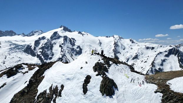 Aerial view of Pointe Ronde summit with skiers and a summit cross overlooking snow covered peaks and rocky ridges under a clear blue sky in Trient Valais Switzerland.