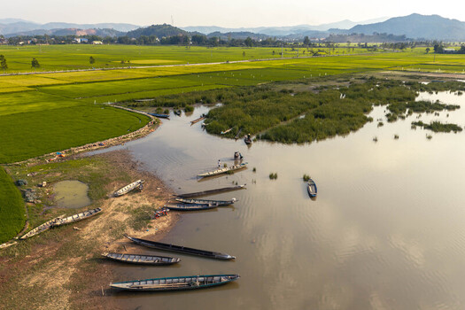 Aerial view of Lak Lake with wooden boats moored along the muddy shore surrounded by vibrant green rice paddies and distant mountains in Lien Son Lak Dak Lak Vietnam. Aerial view of Lak Lake with wooden boats moored along the muddy shore surrounded by vibrant green rice paddies and distant mountains in Lien Son Lak Dak Lak Vietnam.