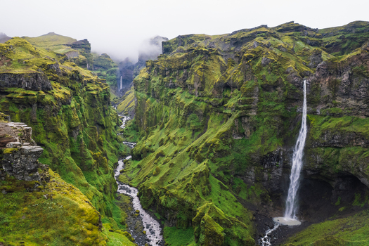 Aerial view of Mulagljufur Canyon with lush green mossy cliffs a winding river and tall waterfalls under a misty sky Eastern Region Iceland. Aerial view of Mulagljufur Canyon with lush green mossy cliffs a winding river and tall waterfalls under a misty sky Eastern Region Iceland.