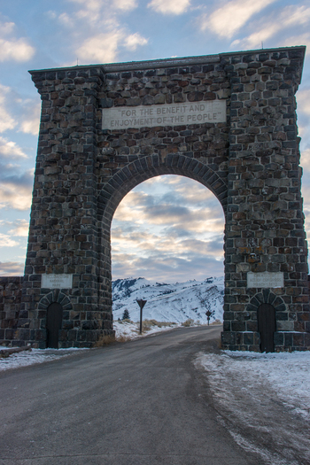 View of the historic stone Roosevelt Arch at the entrance to Yellowstone National Park with snow covered mountains in the background under a cloudy sky United States.