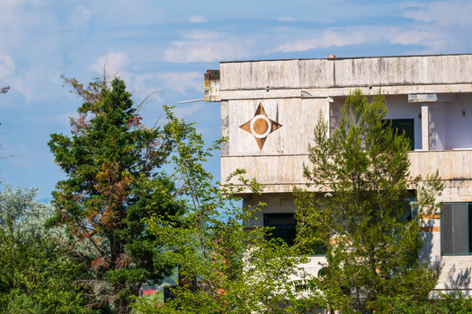 View of a building facade featuring a star shaped decorative emblem surrounded by lush green trees under a blue sky in Divjake Fier County Albania.