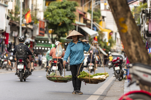 Hanoi Vietnam 11 November 2025 View of a street vendor in a conical hat carrying baskets of fresh vegetables on a shoulder pole while walking through a busy street with motorbikes.