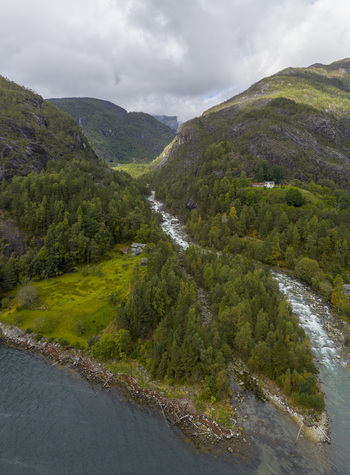 Aerial view of a winding river flowing through a lush green valley surrounded by steep forested mountains and small houses under a cloudy sky Fjaera Vestland Norway. Aerial view of a winding river flowing through a lush green valley surrounded by steep forested mountains and small houses under a cloudy sky Fjaera Vestland Norway.