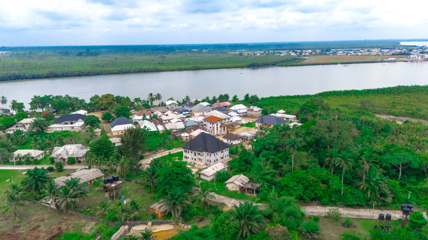 Aerial view of a small settlement with houses nestled among lush tropical vegetation and palm trees along the banks of a wide river in Minima Rivers Nigeria.