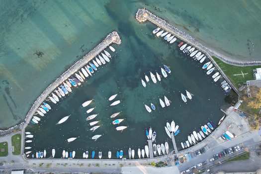 Aerial view of the marina featuring numerous sailboats moored in clear turquoise water along stone breakwaters and a waterfront road Morges Vaud Switzerland. Aerial view of the marina featuring numerous sailboats moored in clear turquoise water along stone breakwaters and a waterfront road Morges Vaud Switzerland.