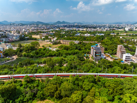 Aerial view of a passenger train traveling through a lush green landscape with dense trees and urban buildings in the background in Udaipur Rajasthan India. Aerial view of a passenger train traveling through a lush green landscape with dense trees and urban buildings in the background in Udaipur Rajasthan India.