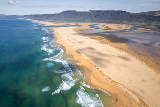 Aerial view of Raudisandur beach featuring golden sand turquoise ocean waves and dark mountain cliffs under a blue sky in Vesturbyggd Iceland. Aerial view of Raudisandur beach featuring golden sand turquoise ocean waves and dark mountain cliffs under a blue sky in Vesturbyggd Iceland.