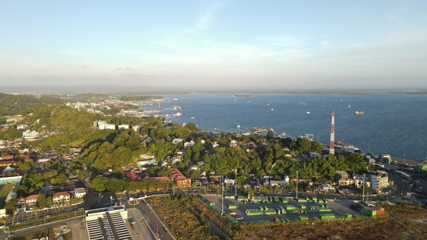 Aerial view of the coastal city of Sorong featuring a busy port with shipping containers a tall industrial chimney and lush green hills in Sorong Southwest Papua Indonesia. Aerial view of the coastal city of Sorong featuring a busy port with shipping containers a tall industrial chimney and lush green hills in Sorong Southwest Papua Indonesia.