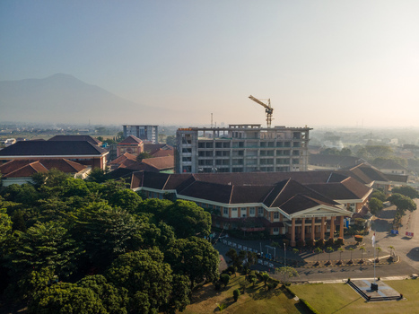 Aerial view of a building complex under construction with a crane surrounded by lush trees and a distant mountain in Banyumas Regency Central Java Indonesia.