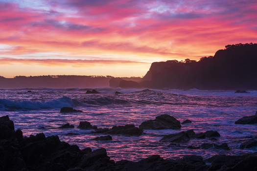 View of Playa de Portizuelo with waves crashing against dark rocks under a vibrant pink and orange sunset sky Spain. View of Playa de Portizuelo with waves crashing against dark rocks under a vibrant pink and orange sunset sky Spain.