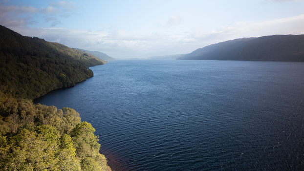 Aerial view of Loch Ness with its deep blue waters surrounded by lush green hills and dense forests under a bright sky in Highland Council Scotland United Kingdom.