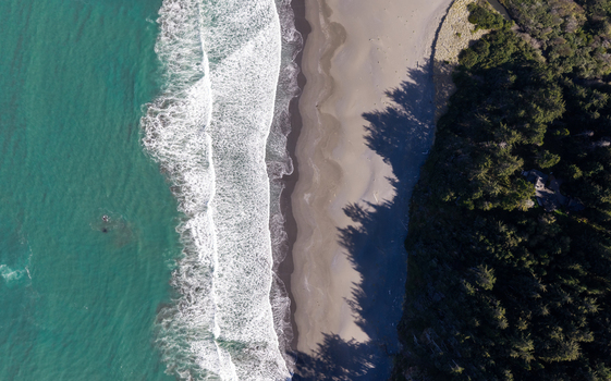 Aerial view of the southern Oregon coast featuring turquoise ocean waves crashing onto a sandy beach bordered by dense evergreen forests in Gold Beach Oregon United States. Aerial view of the southern Oregon coast featuring turquoise ocean waves crashing onto a sandy beach bordered by dense evergreen forests in Gold Beach Oregon United States.