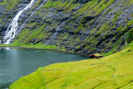 View of a traditional cabin with a grass roof nestled on a green hillside beside a lake with a steep rocky cliff and a waterfall in the background in the Faroe Islands.