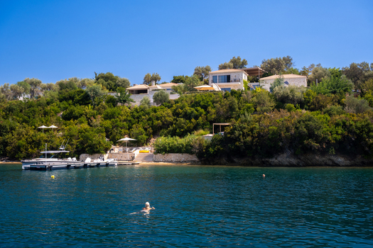 View of a luxury villa nestled on a lush green hillside overlooking the clear blue sea with a person swimming in the foreground in Lefkada Greece.