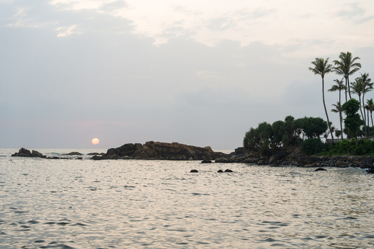View of the sun setting over the Indian Ocean with rocky outcrops and palm trees along the coastline in Galle Southern Province Sri Lanka.