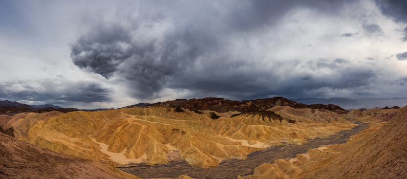 View of the rugged golden brown badlands of Death Valley National Park under a dramatic sky filled with dark storm clouds in the United States.