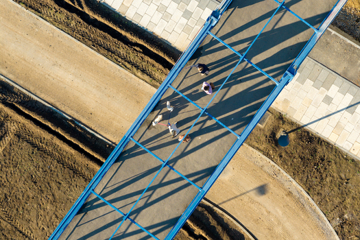 Aerial view of a blue pedestrian bridge with people walking across it casting long shadows over the surrounding dirt paths and paved walkways in Belgrade City of Belgrade Serbia. Aerial view of a blue pedestrian bridge with people walking across it casting long shadows over the surrounding dirt paths and paved walkways in Belgrade City of Belgrade Serbia.