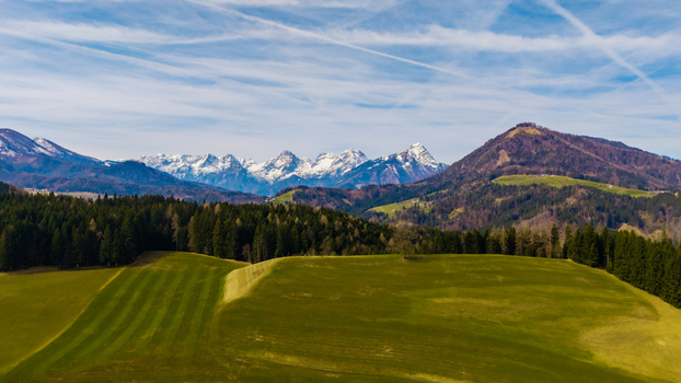 Aerial view of lush green fields meet dark forests leading to snow capped mountains under a vast blue sky Windischgarsten Upper Austria Austria. Aerial view of lush green fields meet dark forests leading to snow capped mountains under a vast blue sky Windischgarsten Upper Austria Austria.