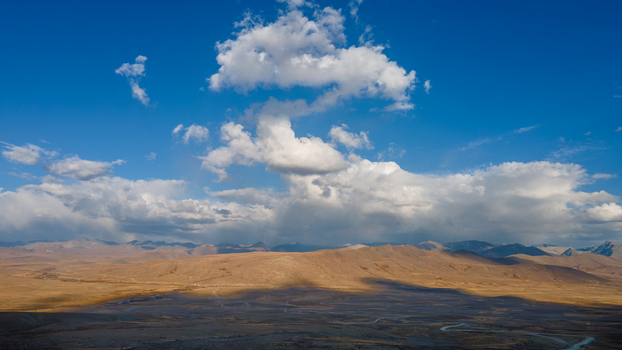 Aerial view of Deosai National Park featuring vast golden plains and rolling mountains under a dramatic blue sky with scattered white clouds in Skardu Gilgit Pakistan.