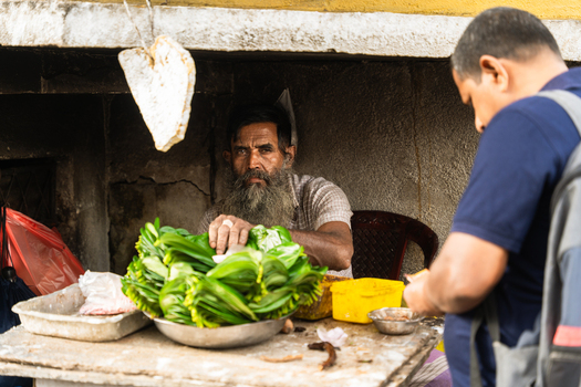 Sri Lanka 20 February 2026 View of a bearded man selling bundles of fresh green betel leaves at a small street stall under a concrete overhang.