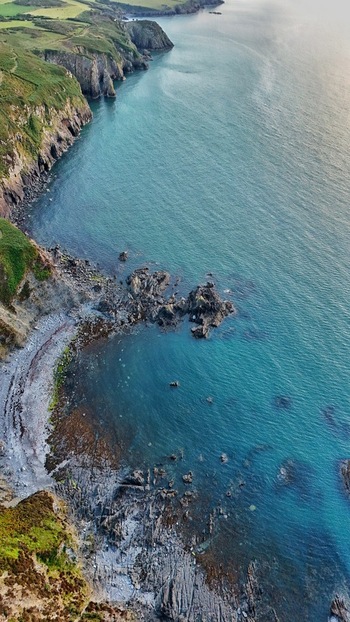 Aerial view of Fishguard Bay featuring steep green cliffs and a rocky shoreline along the turquoise waters of the coast in Fishguard Wales United Kingdom.