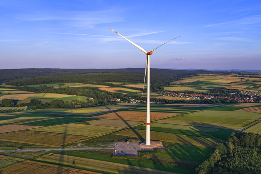 Aerial view of a wind turbine standing in a rural landscape with agricultural fields meadows and a small village in the distance under a clear blue sky in Hofgeismar Hessen Germany.