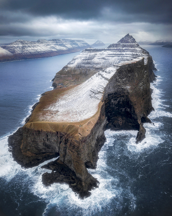 Aerial view of the snow dusted Kallur cliffs and steep rocky ridges surrounded by the dark blue ocean under a moody sky in Klaksvik Northern Isles Faroe Islands. Aerial view of the snow dusted Kallur cliffs and steep rocky ridges surrounded by the dark blue ocean under a moody sky in Klaksvik Northern Isles Faroe Islands.
