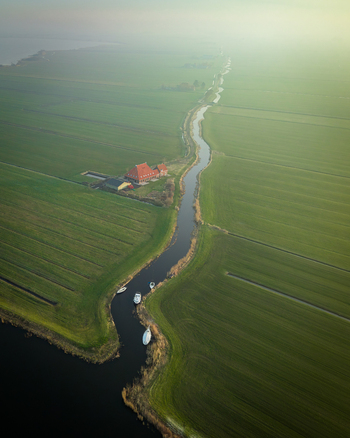 Aerial view of a winding canal through green polders with a farmhouse and moored boats under a misty morning sky Sloten Friesland Netherlands. Aerial view of a winding canal through green polders with a farmhouse and moored boats under a misty morning sky Sloten Friesland Netherlands.