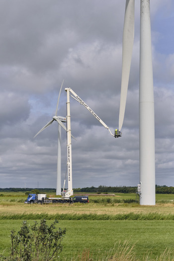 Esbjerg Denmark 24 July 2026 View of crew performing wind turbine blade maintenance using a high reaching cherry picker in a green field under a cloudy sky.