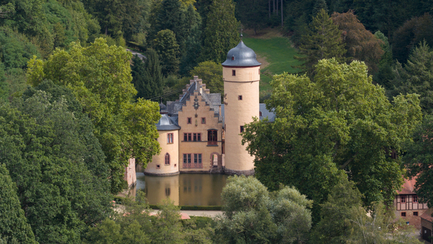 Aerial view of Schloss Mespelbrunn a historic moated castle nestled in a lush forest valley with stone walls towers and surrounding trees in Mespelbrunn Bayern Germany.