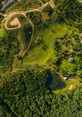 Aerial view of a rural landscape featuring a small pond dense forest and a winding dirt path on a grassy hill in Latvia. Aerial view of a rural landscape featuring a small pond dense forest and a winding dirt path on a grassy hill in Latvia.