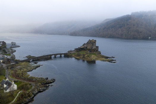 Aerial view of Eilean Donan Castle on its small island connected by a stone bridge over the calm waters of Loch Duich under misty hills Dornie Scotland United Kingdom.