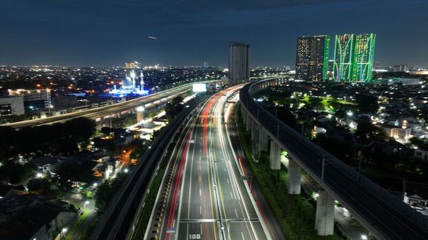 Aerial view of long exposure light trails on a highway and elevated railway tracks next to illuminated skyscrapers at night in Bekasi Barat West Java Indonesia.