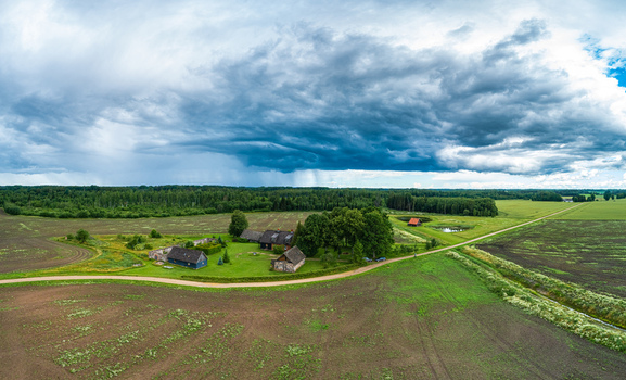 Aerial view of a rural farmstead surrounded by vast agricultural fields and lush trees under a dramatic dark storm sky in Latvia. Aerial view of a rural farmstead surrounded by vast agricultural fields and lush trees under a dramatic dark storm sky in Latvia.