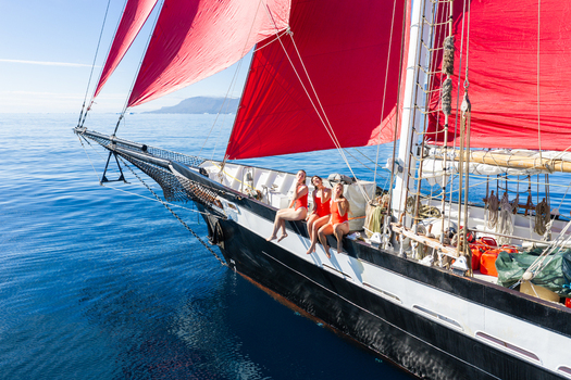 Greenland 30 June 2026 Aerial view of a sailing ship with striking red sails gliding over deep blue water featuring three women in red swimsuits sitting on the bow. Greenland 30 June 2026 Aerial view of a sailing ship with striking red sails gliding over deep blue water featuring three women in red swimsuits sitting on the bow.